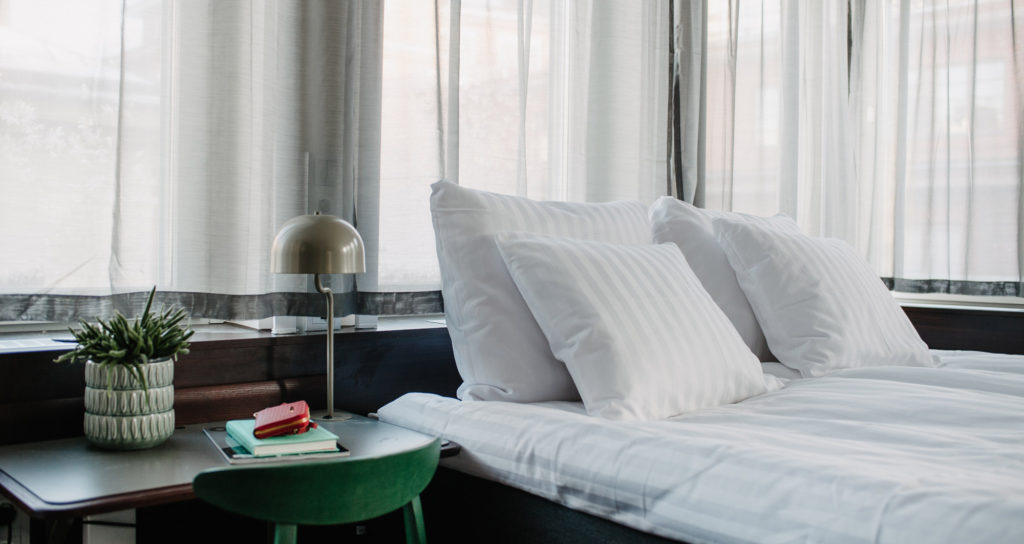 Del av säng med kuddar, skrivbord och sänglampa. Boka hotellrum i Stockholm